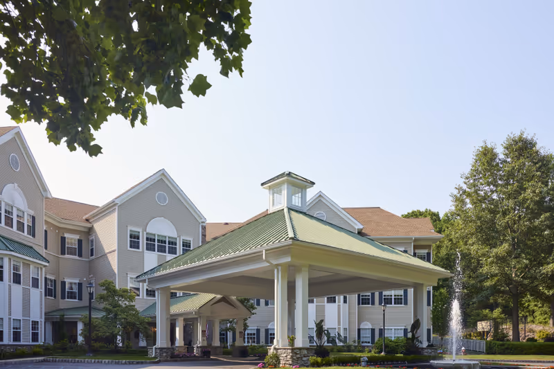 Exterior view of Brandywine Serenade by Monarch senior living facility showing a large covered entrance with a green roof, beige building with multiple windows, trees, and a water fountain in front.