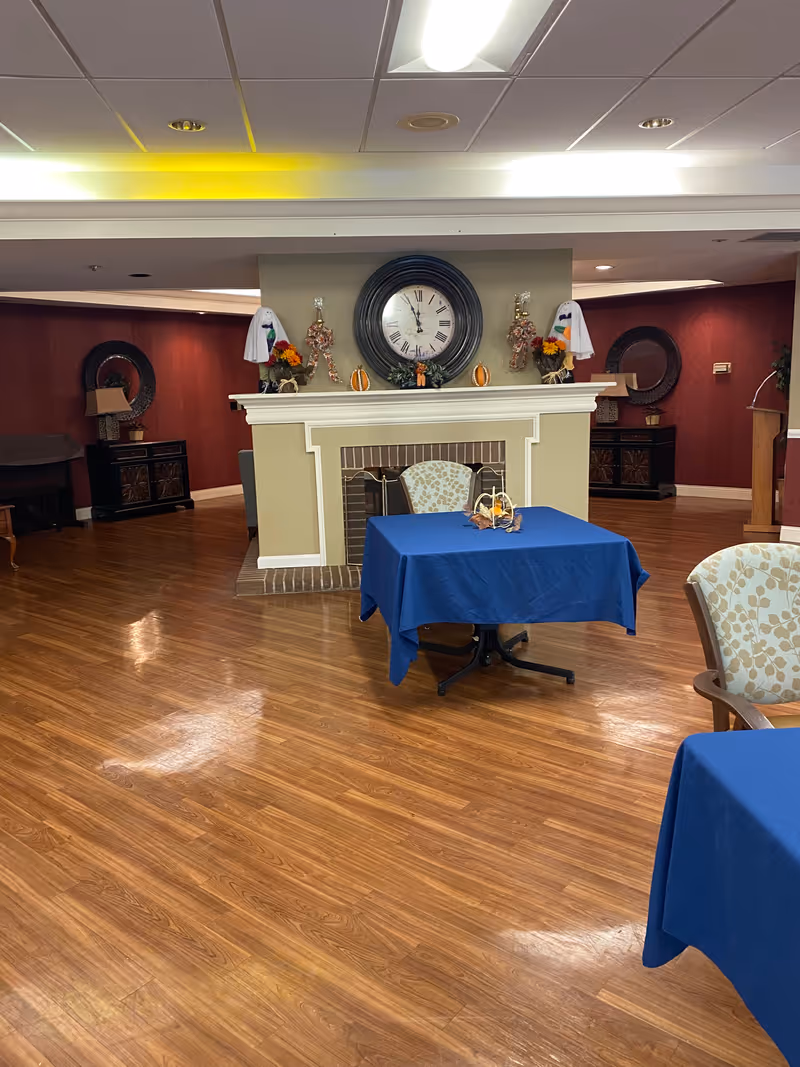 Interior view of a common area with wooden flooring, a fireplace decorated with autumn-themed items and a large round clock above it. There are tables covered with blue tablecloths and chairs with patterned upholstery. The walls are painted in warm tones with mirrors and lamps on side tables.