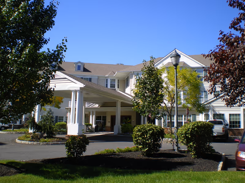 Exterior view of a senior living facility building with white siding and a covered entrance supported by white columns. There are trees, bushes, and a lamp post in the landscaped front area, with a clear blue sky above.