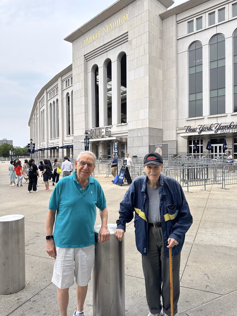 Two men standing on a plaza in front of Yankee Stadium Gate 6 with crowds, barriers, and metal bollards visible.