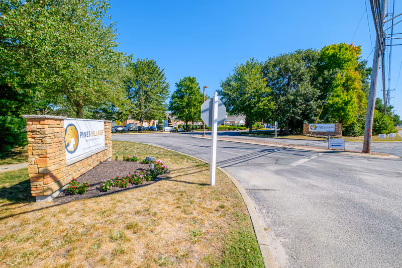 Entrance to Pines Village senior living community showing stone signage, a driveway, and surrounding trees.