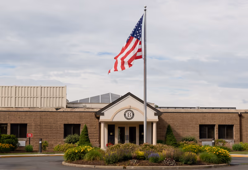 Front exterior view of Bridgeway Care and Rehabilitation Center at Bridgewater, featuring a single-story brick building with a central entrance topped by a triangular pediment. An American flag flies on a flagpole in front of the entrance, surrounded by landscaped bushes and flowers.