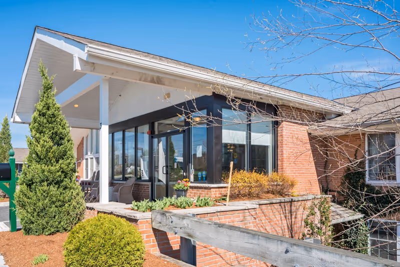 Exterior view of Bentley Assisted Living at Branchville showing a brick building with large windows and a covered entrance. There are green shrubs and small trees in the landscaped area around the building under a clear blue sky.