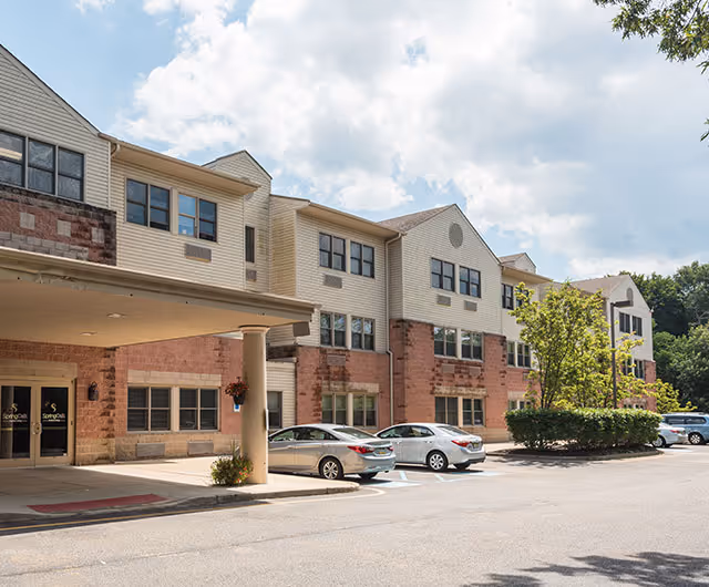 Exterior view of a multi-story senior living facility building with a covered entrance, several parked cars, and trees under a partly cloudy sky.