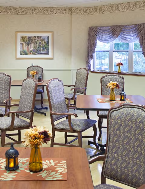 Interior view of a dining room in a senior living facility with multiple wooden tables and cushioned chairs arranged neatly. Each table has a small vase with flowers and a decorative lantern. A window with purple drapes allows natural light to enter the room, and a framed painting hangs on the wall.