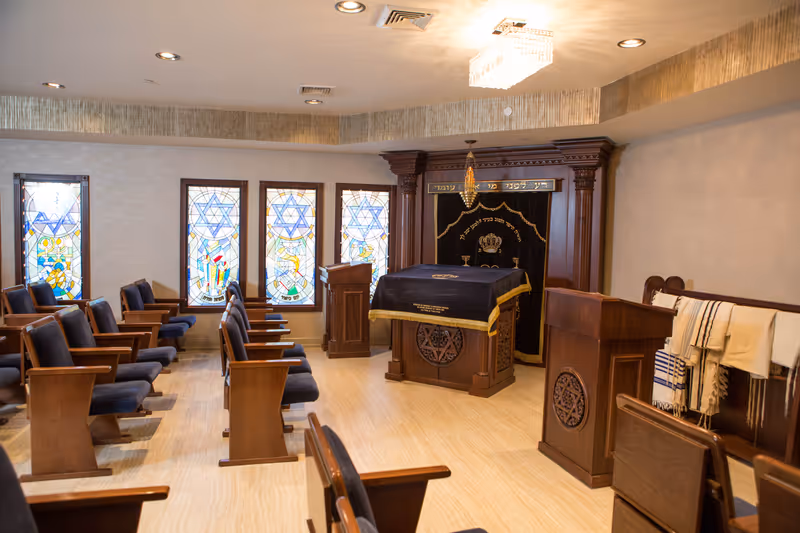 Interior view of a small synagogue or prayer room with wooden pews and chairs arranged facing a central podium and an ark covered with a black cloth featuring a crown emblem. The room has stained glass windows with Star of David designs and religious symbols, a chandelier on the ceiling, and prayer shawls hanging on a wooden rack.