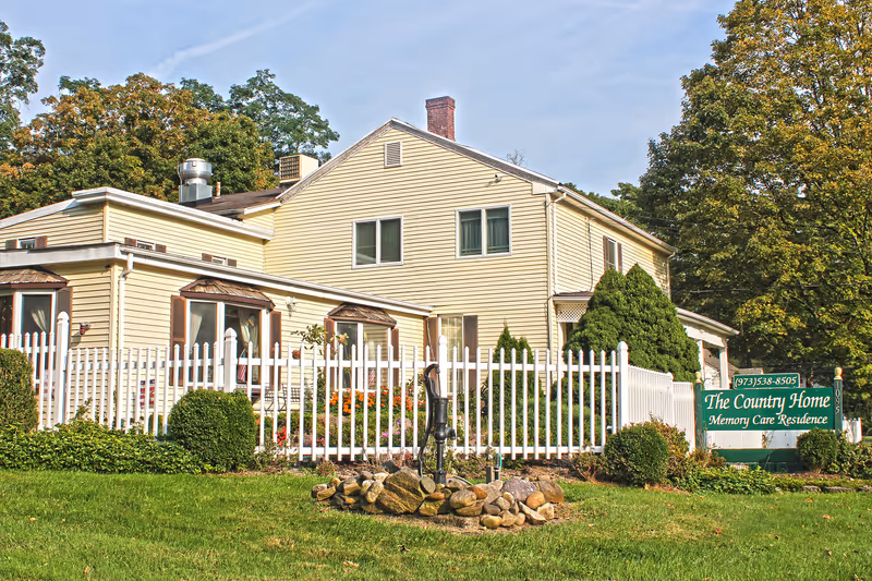 Exterior view of The Country Home Memory Care Residence, a two-story beige building with white trim surrounded by a white picket fence, green shrubs, and a small garden with flowers. A green sign with white text displays the facility name and phone number. Trees are visible in the background under a clear sky.