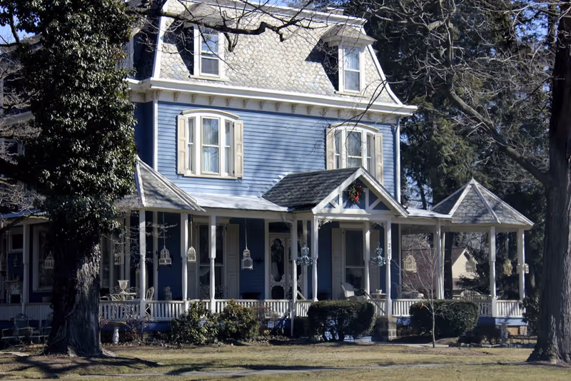 A large, two-story blue house with white trim and a wraparound porch. The house has multiple windows with shutters, a steep roof with dormer windows, and is surrounded by trees and bushes. The porch is decorated with hanging lanterns and has seating areas.