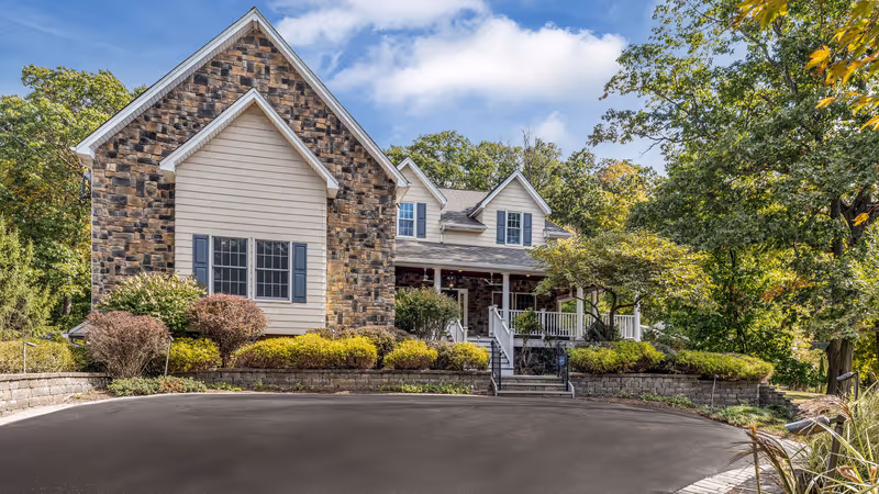 Front exterior view of a senior living facility named Clover Hill Senior Living, featuring a stone and siding facade, multiple windows, a covered porch with white railings, surrounded by landscaped bushes and trees under a partly cloudy sky.