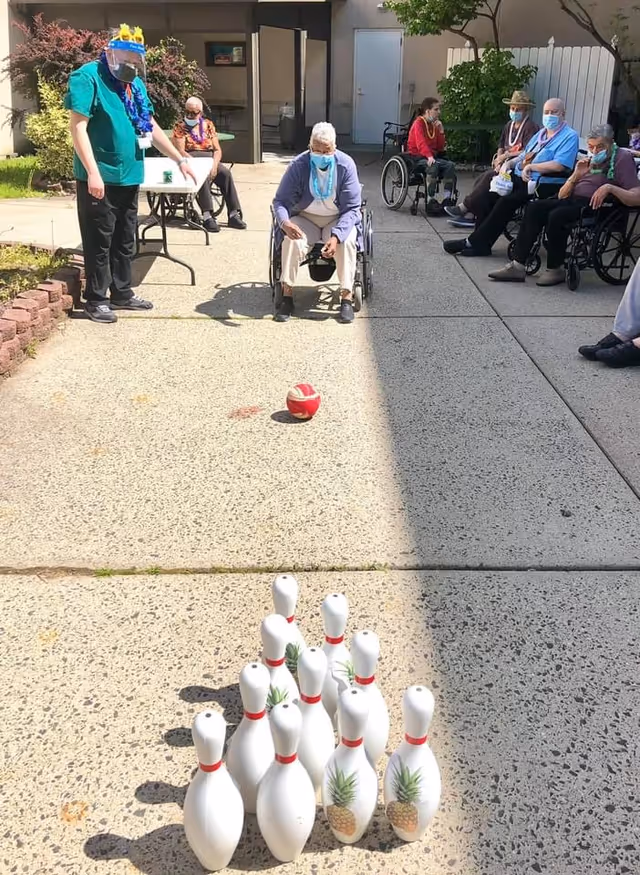 A group of elderly individuals, some in wheelchairs, are playing an outdoor bowling game on a paved area. A staff member wearing a face shield and mask is standing nearby, observing and assisting. The bowling pins have pineapple designs on them, and one person in a wheelchair is rolling a red ball towards the pins. The setting appears to be a courtyard or patio area with some greenery and a building entrance in the background.
