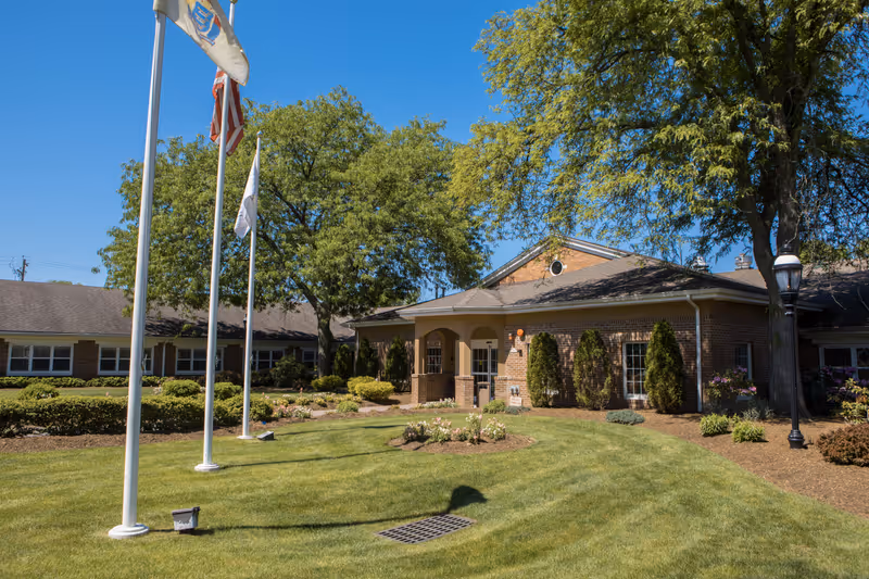 Exterior view of a senior living facility building with a well-maintained lawn, flower beds, and three flagpoles displaying flags. The building is made of brick with large windows and surrounded by trees under a clear blue sky.