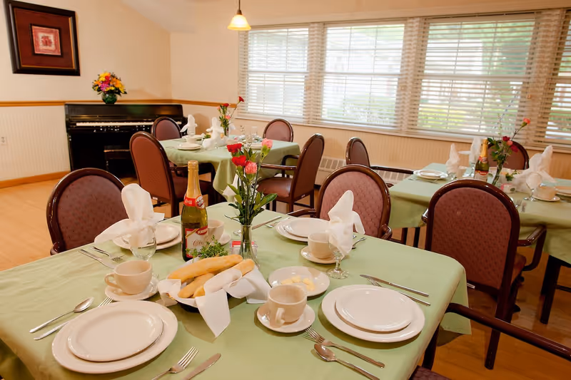 A dining room set up with tables covered in green tablecloths, each set with white plates, cups, silverware, and folded white napkins. Breadsticks in a basket, small plates with butter, and bottles of sparkling cider are on the tables. There are chairs with maroon upholstery around the tables, a piano against the wall, a framed picture, and large windows with blinds letting in natural light.