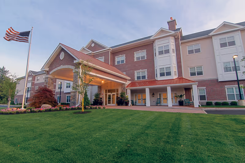 Exterior view of a three-story senior living facility building with a covered entrance, well-maintained green lawn, flower beds, and an American flag on a flagpole.