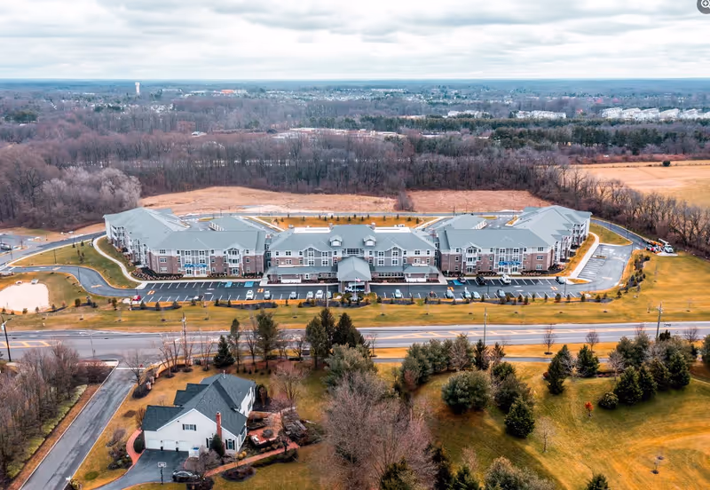 Aerial view of Parkers Bend Retirement Community showing a large, U-shaped building with a gray roof and brick facade surrounded by parking lots, roads, and landscaped green areas with trees and grass. The background includes a forested area and a cloudy sky.
