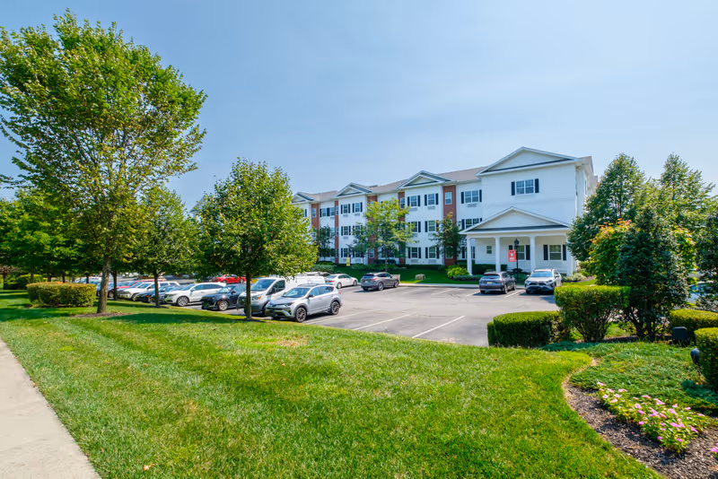 Exterior view of a senior living facility building with white siding and brick accents, surrounded by green trees and a well-maintained lawn. There is a parking lot with several cars parked in front of the building under a clear blue sky.