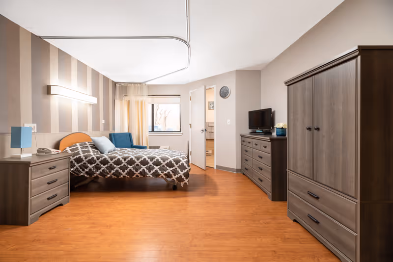 A well-lit bedroom in a senior living facility featuring a bed with patterned bedding, a blue armchair near a window with curtains, a nightstand with a blue lamp and telephone, a dresser with a small TV and decorative flowers, and a large wardrobe. The room has wood flooring and striped wallpaper on one wall.