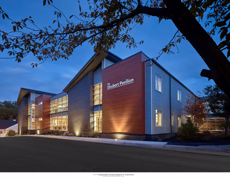 Exterior view of The Shubert Pavilion building at dusk with modern architecture featuring large windows and a combination of wood and metal siding, framed by tree branches in the foreground.