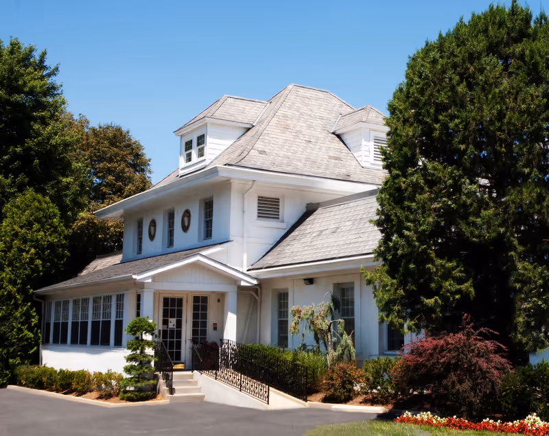 White two-story building with a porch entrance, surrounded by trees and landscaping under a clear blue sky.