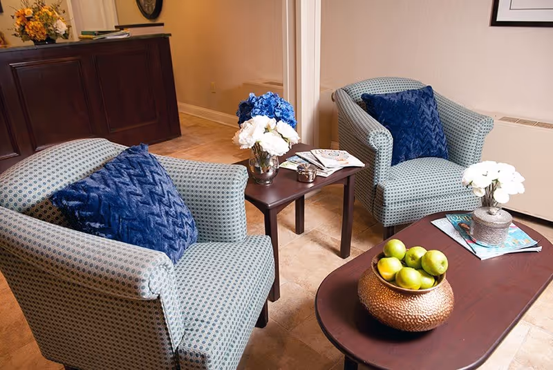 A cozy seating area with two patterned armchairs each with a blue cushion, a small wooden side table with a vase of blue and white flowers and magazines, and a wooden coffee table with a bowl of green apples and a small flower pot with white flowers. The floor is tiled and there is a dark wooden reception desk in the background.