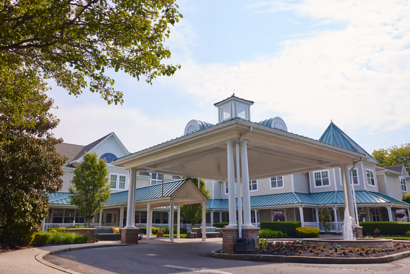 Exterior view of Brandywine Brandall Estates by Monarch, showing a large covered entrance with white columns and a landscaped roundabout with a fountain. The building has light-colored siding and teal metal roofing, surrounded by trees and shrubs under a partly cloudy sky.