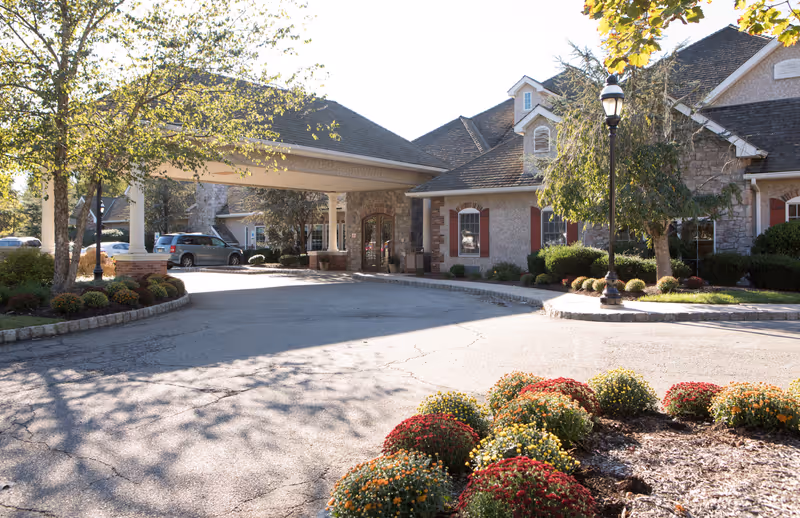 Exterior view of CareOne at Livingston facility showing a driveway with a covered entrance supported by white columns, a stone and stucco building with red window shutters, landscaped flower beds with colorful flowers, trees, and a black street lamp.