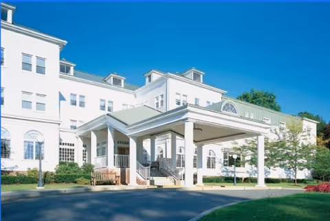 Exterior view of a large white senior living facility building with multiple windows, a covered entrance with columns, and a driveway in front under a clear blue sky.