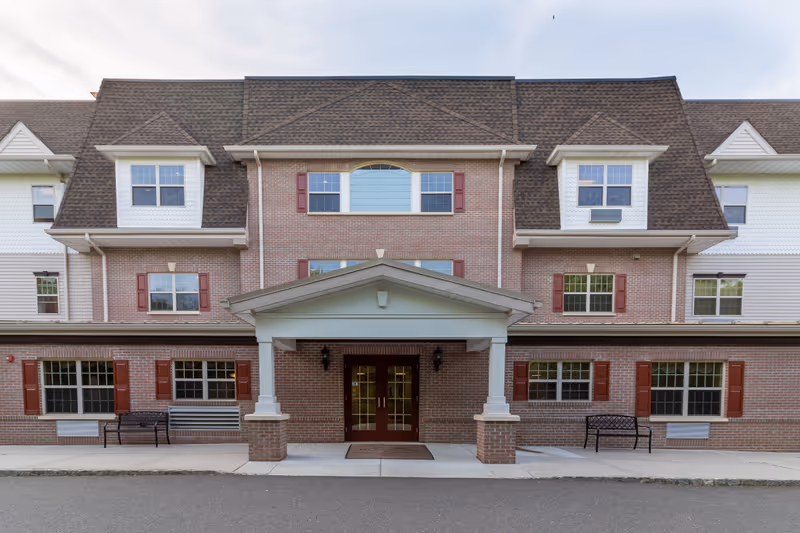 Front exterior view of a multi-story brick building with white trim and red shutters. The entrance has a covered porch with two white columns and double glass doors. There are benches on either side of the entrance on the sidewalk.