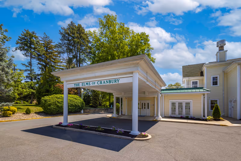 Entrance of The Elms of Cranbury facility with a covered drop-off area supported by white columns, surrounded by greenery and trees under a partly cloudy blue sky.