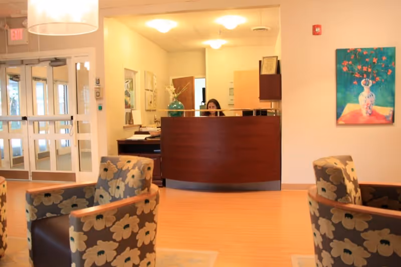 Reception area of a senior living facility with a wooden front desk where a woman is seated. The room has light-colored walls, a painting of a vase with flowers on the right wall, and floral-patterned armchairs in the foreground. Double glass doors are visible on the left side, allowing natural light to enter.