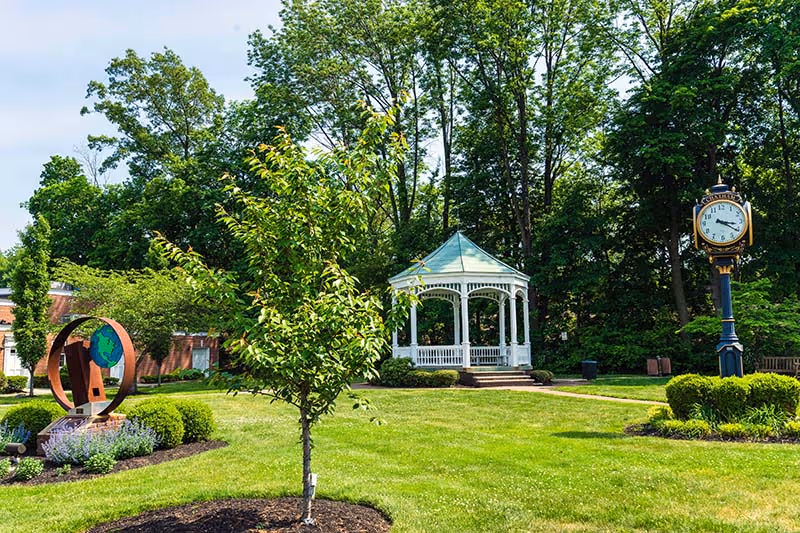 A well-maintained outdoor garden area with a small tree in the foreground, a white gazebo with a green roof in the middle, a decorative clock on a post to the right, and a modern sculpture on the left, surrounded by green grass and trees.