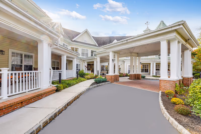 Covered porte-cochere and front entrance of a light-colored senior living building with white columns, a porch, and landscaped walkway.