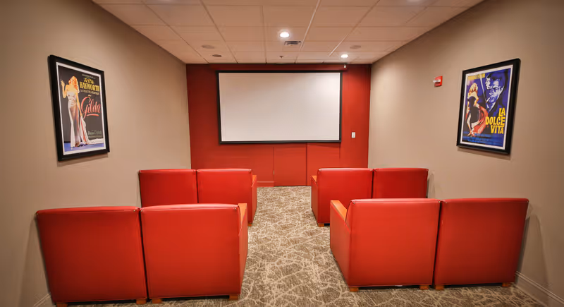 Small theater room with six red armchairs arranged in two rows facing a blank projector screen mounted on a red accent wall. The room has beige walls with framed vintage movie posters on each side and a patterned carpet floor.