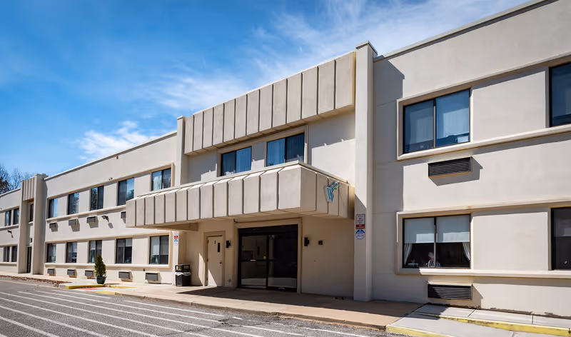 Exterior view of a two-story beige building under a clear blue sky, featuring multiple windows and a covered entrance with a small sign displaying a 'W'. The building is part of Willow Springs Rehabilitation & Healthcare Center.