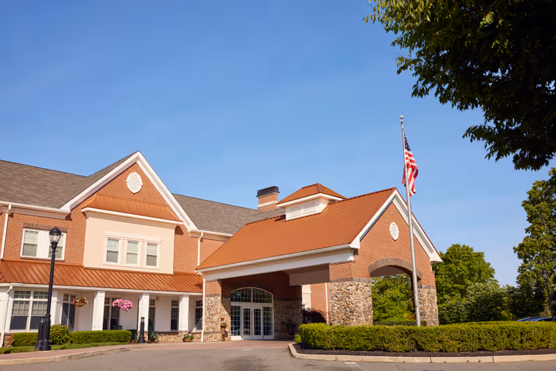 Exterior view of a senior living facility building with a brick facade, stone pillars, and a covered entrance. There is an American flag on a flagpole near the entrance, and the sky is clear and blue. Trees and shrubs surround the building.