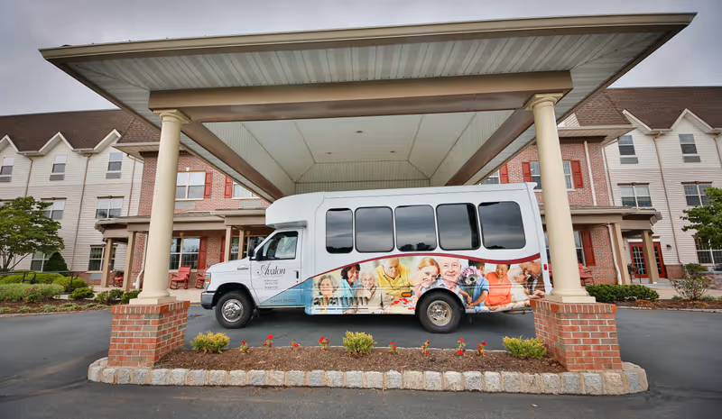 A white shuttle bus with images of smiling elderly people and a dog parked under a large covered entrance of a multi-story assisted living facility with brick and siding exterior and red shutters.