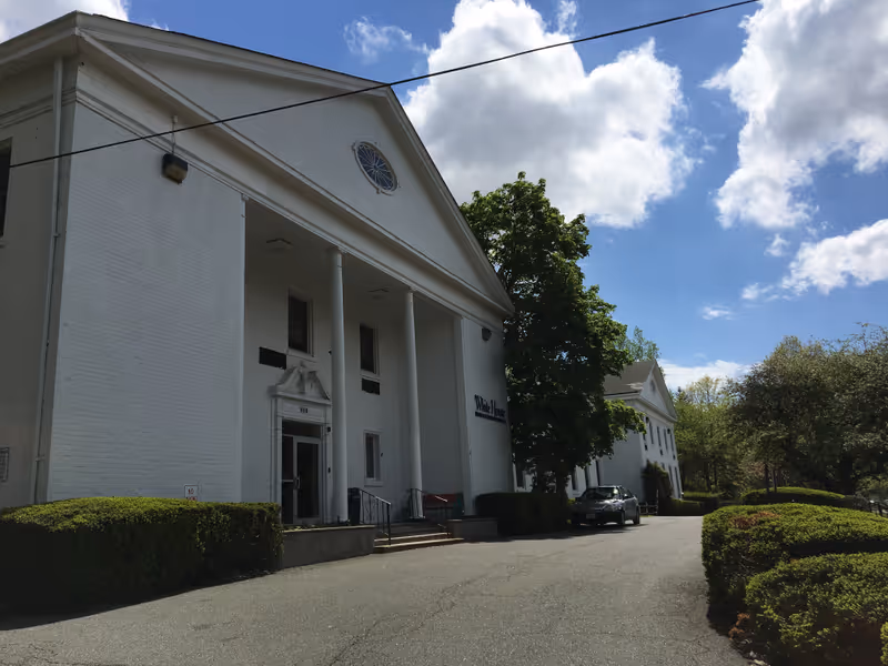 Exterior view of a large white building with classical architecture featuring tall columns and a triangular pediment. The building is surrounded by greenery, including bushes and trees, under a partly cloudy blue sky. A car is parked near the entrance.