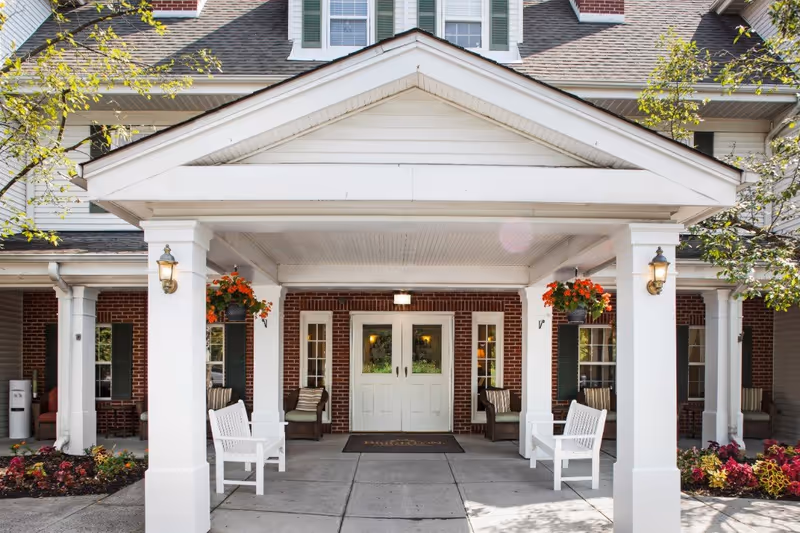 Entrance of a senior living facility with a covered porch supported by white columns, two white benches, hanging flower baskets with orange flowers, and a brick building facade with double doors and windows.