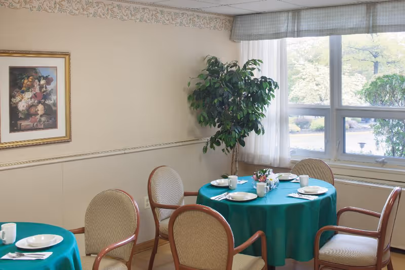 A dining area with round tables covered in green tablecloths, set with white plates, cups, and silverware. Four cushioned chairs surround each table. A large window with sheer curtains lets in natural light, and a potted plant is placed near the window. A framed floral painting hangs on the wall.