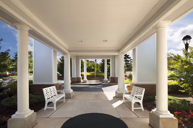 Covered building entrance with white columns, benches, and a circular driveway surrounded by landscaping.