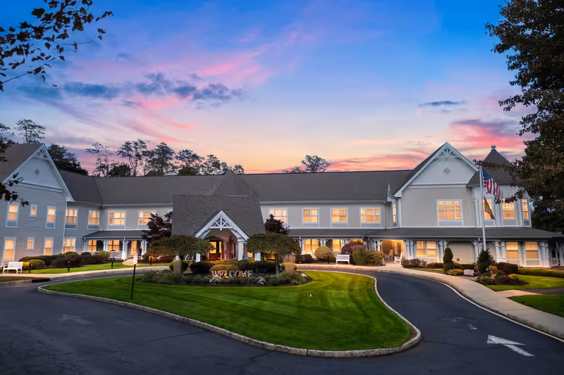Exterior view of a large, two-story senior living facility building at sunset with well-maintained landscaping, a circular driveway, and American flags near the entrance.