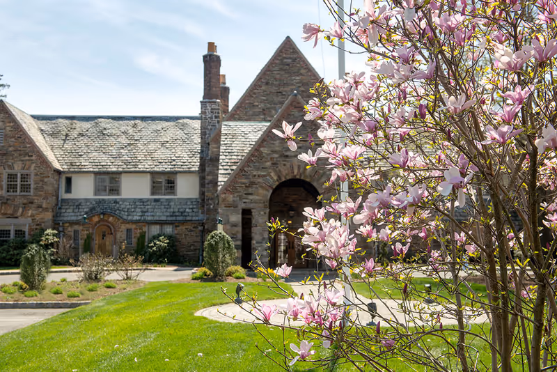 Stone building with a slate roof and arched entrance surrounded by a well-maintained lawn and blooming pink magnolia tree in the foreground under a clear blue sky.