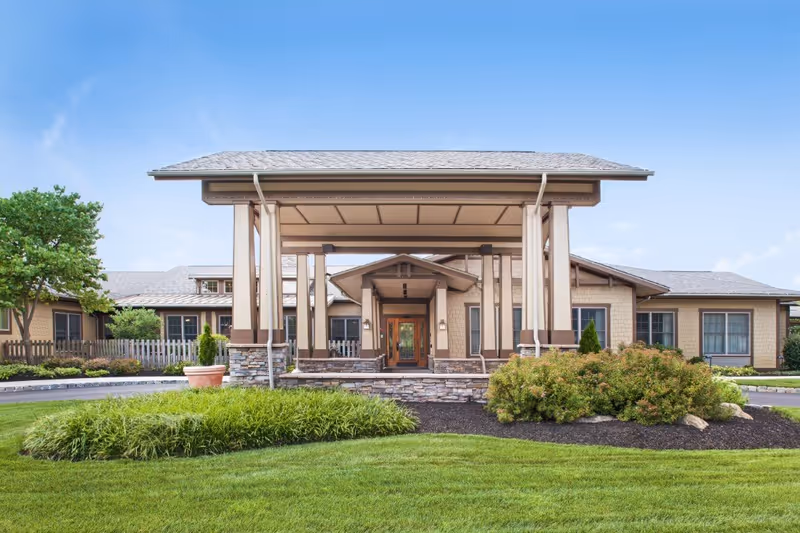 Front exterior view of a single-story senior living facility building with a covered entrance supported by columns, surrounded by well-maintained green grass, bushes, and a tree under a clear blue sky.