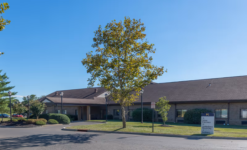 Exterior view of a single-story brick building with a brown roof, surrounded by landscaped bushes and trees under a clear blue sky. A sign near the entrance indicates directions for Main Entrance, Ambulance Receiving, and Delivery.