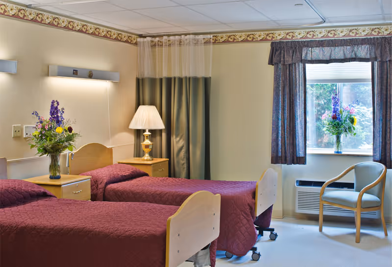 A senior living facility bedroom with two single beds covered in maroon quilts. Each bed has a wooden headboard and is separated by a wooden nightstand with a lamp and a vase of colorful flowers. There is a window with floral curtains letting in natural light, and a wooden chair is placed near the window. The walls are light-colored with a floral border near the ceiling.