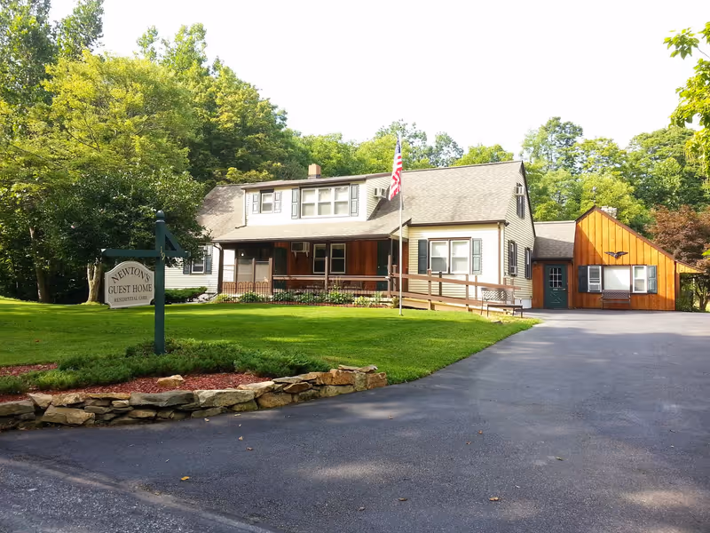 Front exterior of Newton's Guest Home, a two-story house with a lawn, driveway, American flag, and a sign.