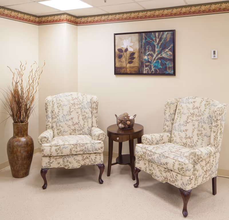 Two floral patterned armchairs with wooden legs placed on either side of a small round wooden table with a decorative bowl on top. A large vase with dried branches is positioned to the left of the chairs. A framed artwork featuring floral and abstract designs hangs on the beige wall behind the chairs. The room has a beige floor and a decorative floral border near the ceiling.