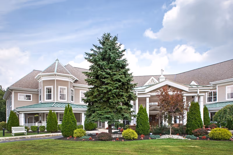 Exterior view of a senior living facility building with a large evergreen tree and landscaped garden in front, featuring a covered entrance with white columns and a green roof under a partly cloudy sky.