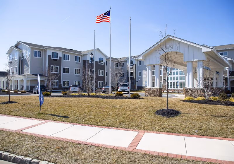 Three-story senior living building with a covered porte-cochere entrance, American flag, and landscaped lawn in front.