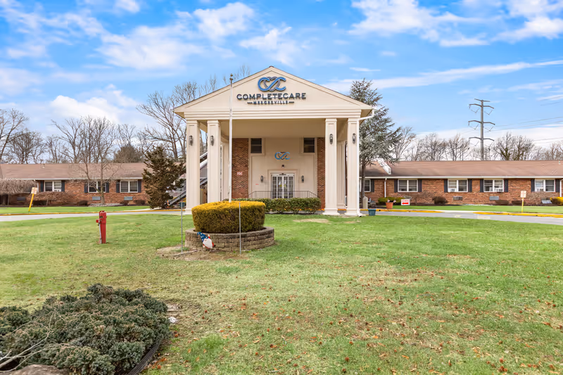 Front exterior view of Complete Care at Mercerville facility showing a building with a large portico supported by four columns, a manicured lawn, and a clear blue sky with some clouds.