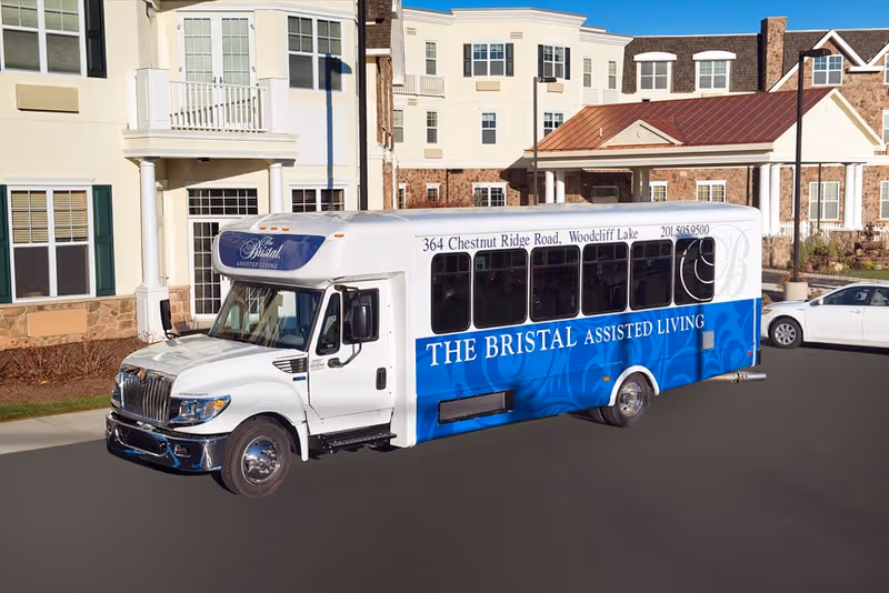 A white and blue shuttle bus parked in front of a large assisted living facility building. The bus has 'THE BRISTAL ASSISTED LIVING' written on its side along with the address '364 Chestnut Ridge Road, Woodcliff Lake' and phone number '201.505.9500'. The building behind has multiple windows, balconies, and a covered entrance.
